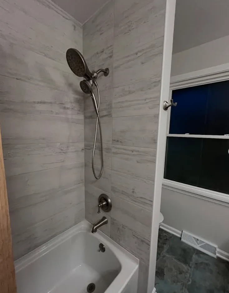 Bathroom tub and shower surround finished in large-format gray wood-grain tile with a brushed nickel shower fixture, handheld sprayer and white alcove tub