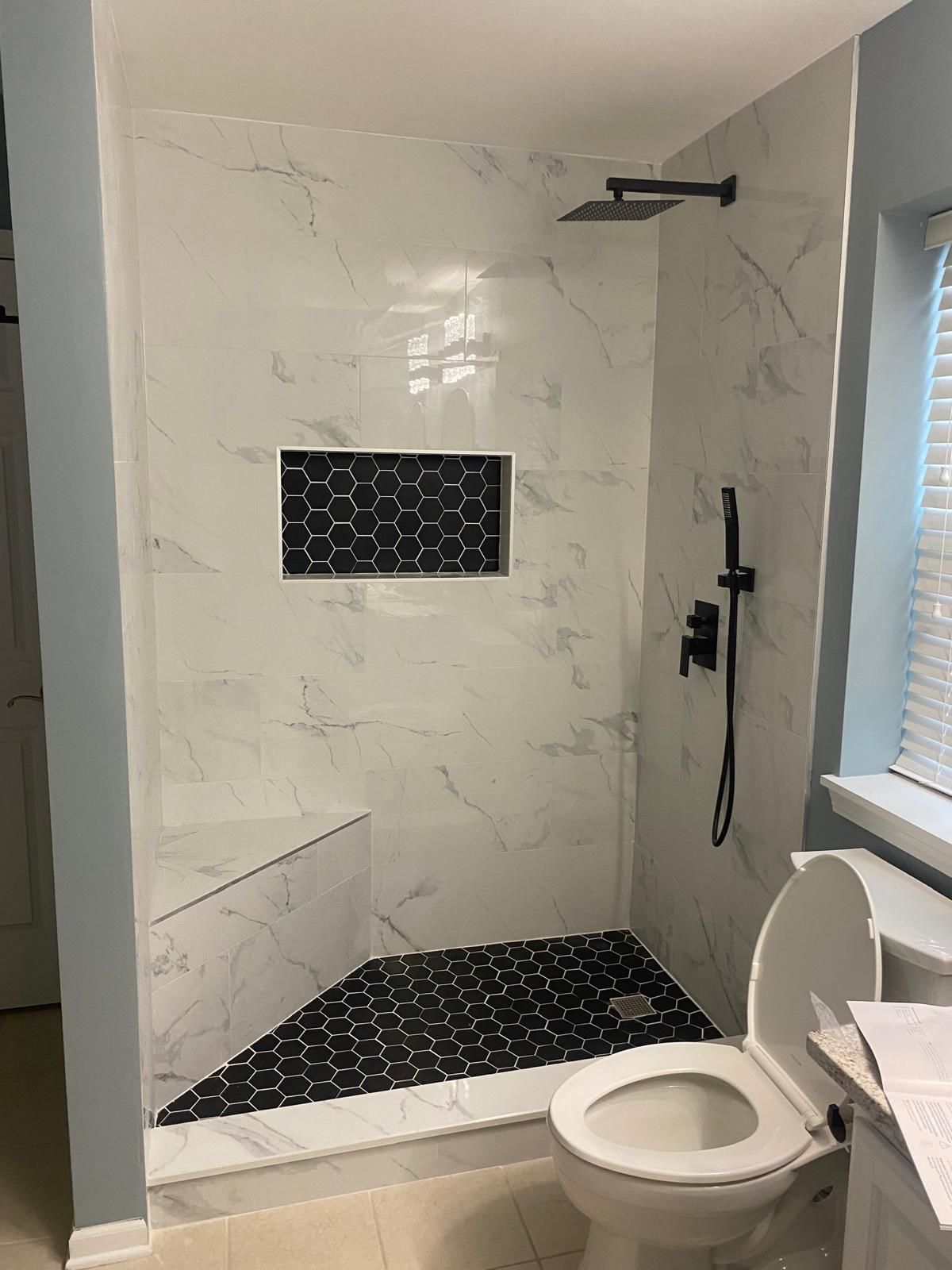 Walk-in shower tiled in large-format white marble-look porcelain with a black hexagon mosaic floor and matching recessed niche, matte black rain shower head and handheld sprayer, and a built-in bench