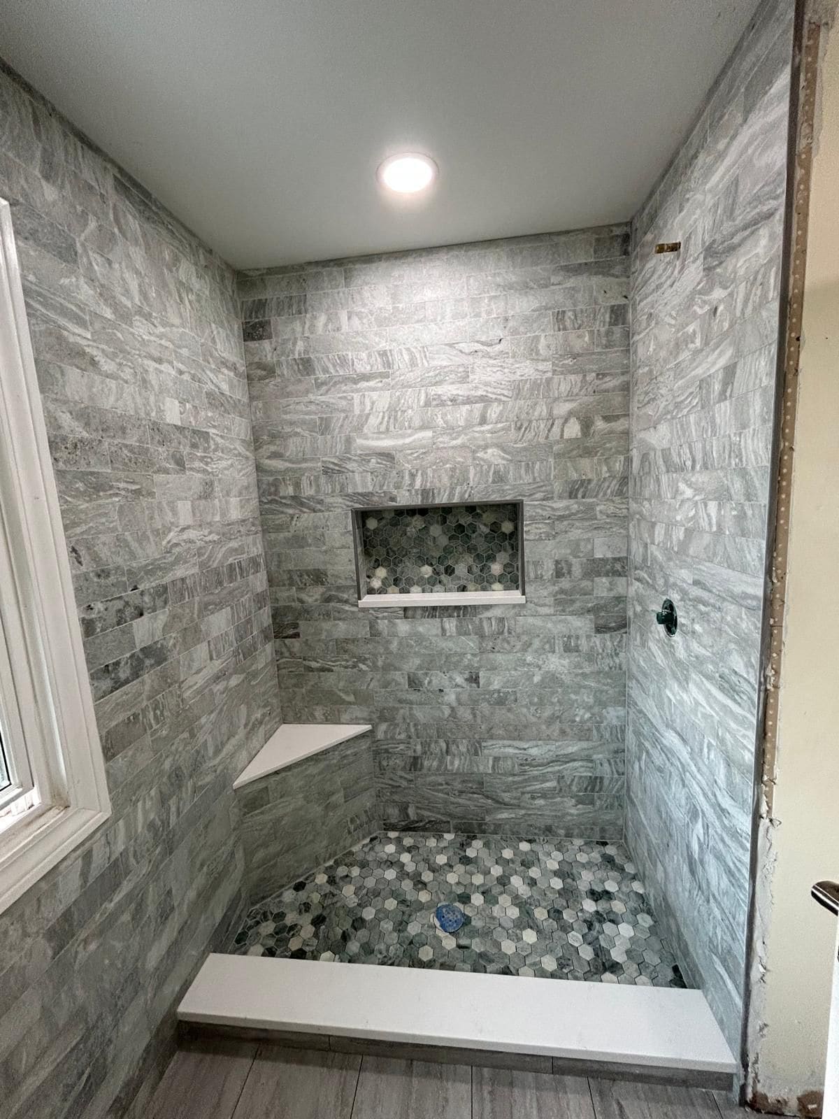 Walk-in shower tiled floor to ceiling in gray marbled stone-look plank tile, with a white corner bench and curb, a recessed niche and shower floor in gray and green hexagon mosaic, and a recessed ceiling light